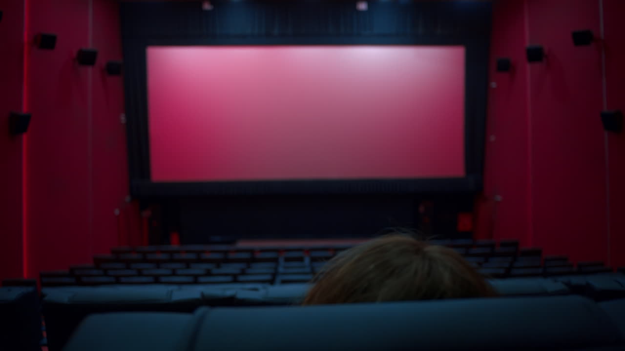 Woman taking her seat in a large empty movie theater auditorium with illumination and blank screen
