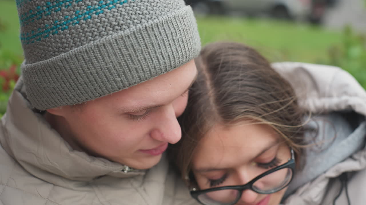 Close up of man wearing knitted cap gently kissing wife on head as she leans into him with eyes closed, surrounded by soft greenery swaying in breeze