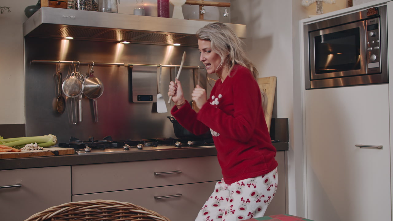 Una giovane donna che canta e balla con un maglione natalizio festivo nella sua cucina, mentre prepara un pasto e si diverte ascoltando musica.