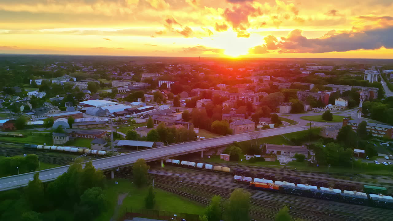 drone aéreo disparado sobre un tren de carga de vagones de petróleo que pasa bajo un puente a través de una ciudad con la puesta de sol en el fondo