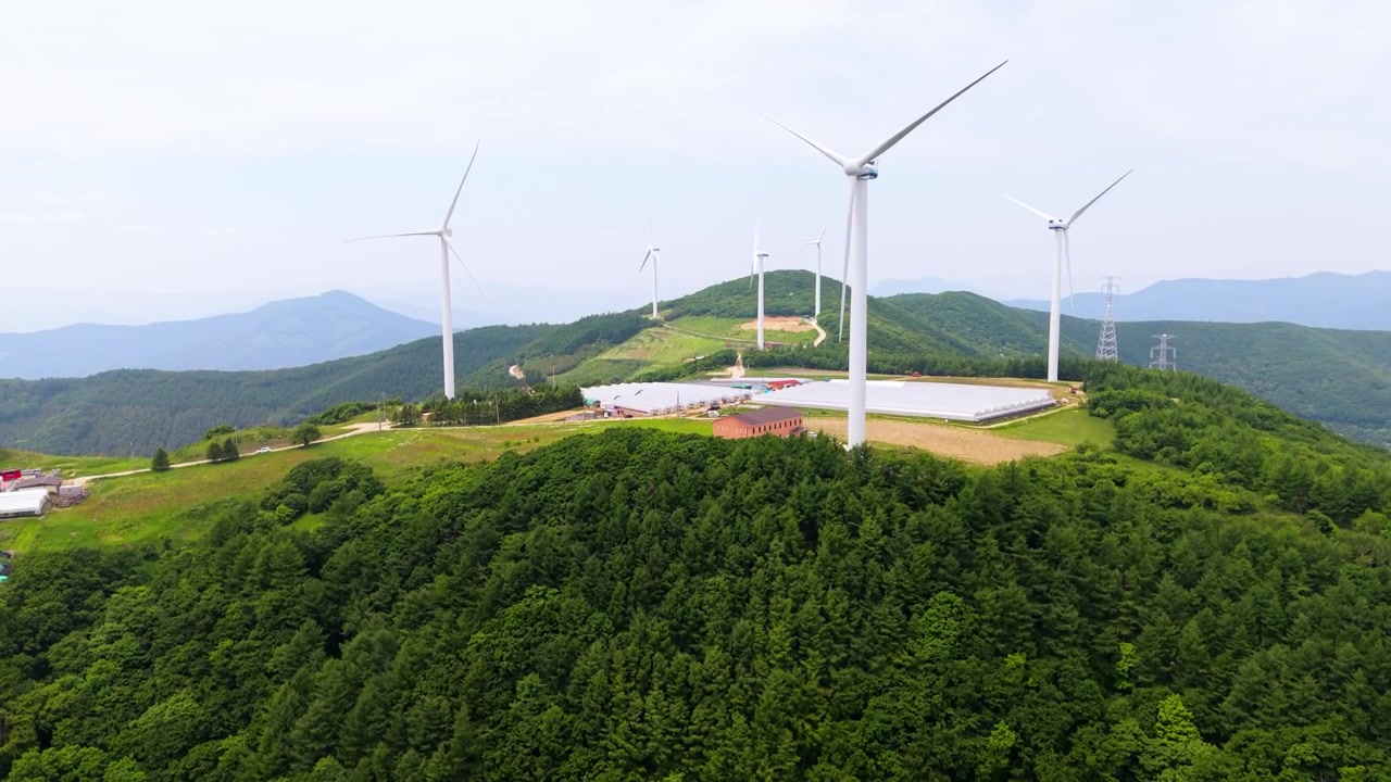 韓国平昌（ピョンチャン）のユクベクマギ牧場にある、森林に覆われた山頂に複数の風力タービンと温室農場が広がる様子をカメラが上昇しながら捉える。