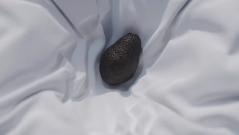 Mesmerizing-slow-motion-footage-of-an-avocado-descending-onto-a-pristine-white-sheet,-followed-by-being-carefully-enfolded,-invoking-a-serene,-minimalist-aesthetic