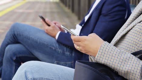 Close Up Of Business Commuters Sitting On Seat On Railway Platform With Mobile Phones