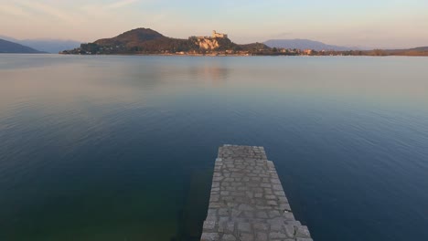 peaceful establishing shot of concrete jetty on lake maggiore calm water in italy, angera castle in background