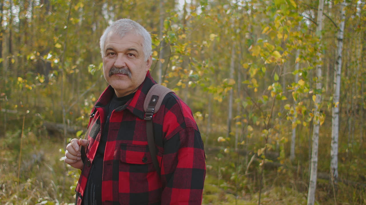 positivo excursionista de mediana edad con mochila está sonriendo y mirando la cámara retrato masculino en el bosque en el día de otoño durante la caminata
