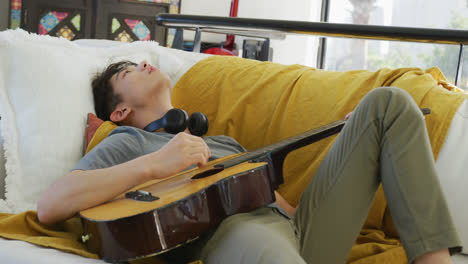 asian male teenager with headphones playing guitar and lying in living room