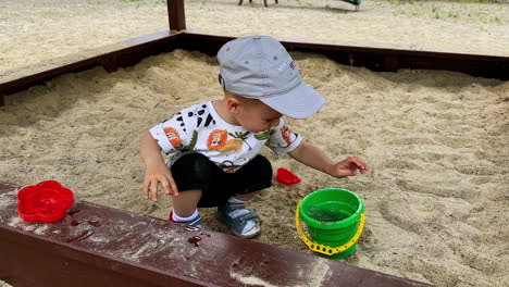 small kid sits in the sandbox playing with his toys. smiling kid splashes water from his toy bucket.