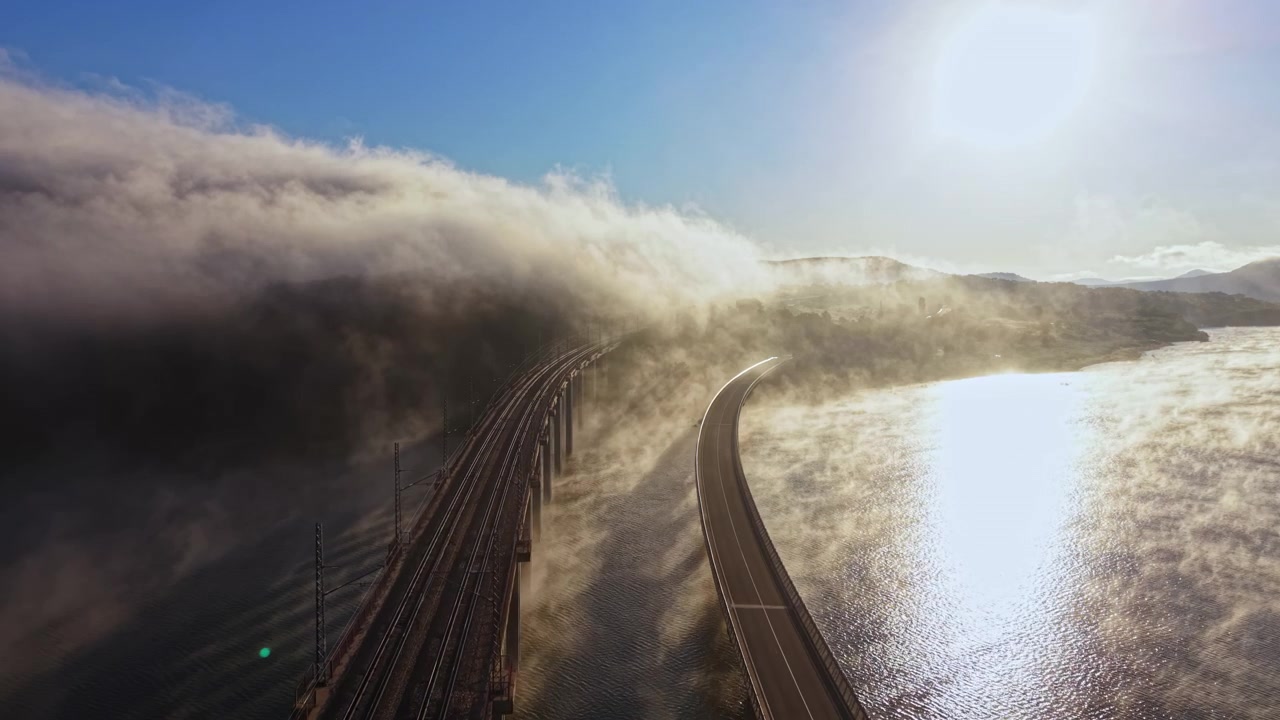 Sabahın erken saatlerinde, sisle kaplı bir manzarada kıvrımlı bir nehir ve tren rayları üzerine süzülen güneş ışığı.