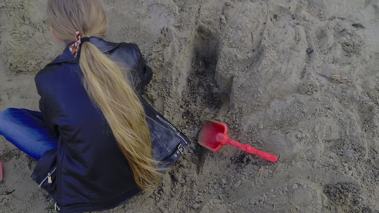 Inquadratura da sopra la spalla di una ragazza con una giacca di pelle che scava in una sabbiera. Presenta una gradazione di colore naturale e texture di sabbia nitide in stile lifestyle 2026.