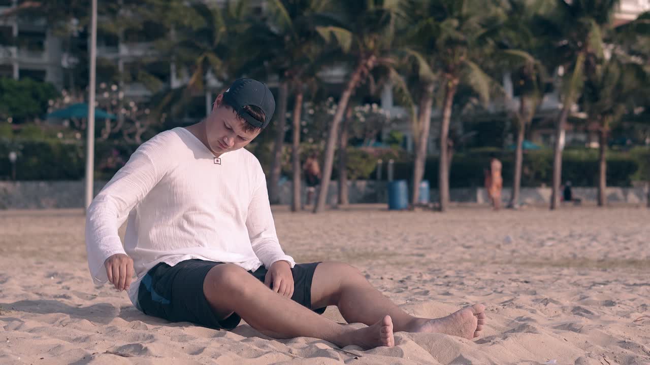 모자를 입은 남자가 일찍 어두워지는 해변에 모래를  ⁇ 고 앉아 있습니다.