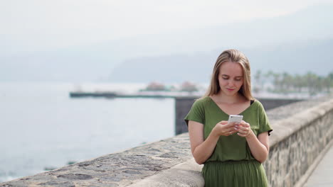 A-traveler-holds-a-mobile-phone-in-her-hand-against-the-backdrop-of-a-beach's-seascape-horizon.-The-tourist-gazes-at-the-sun-kissed-blue-ocean,-embodying-a-vibrant-summer-lifestyle