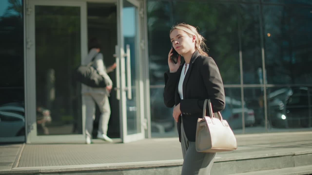 Side view of banker on phone with customer showing frustration during tense conversation while walking past mall with glass facade reflecting cars, trees, and passerby entering entrance