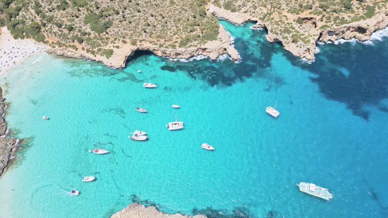 The midday sun shines brightly on Cala Varquas, illuminating the turquoise waters as boats fill the picturesque cove, creating a vibrant scene of summer activity and relaxation.