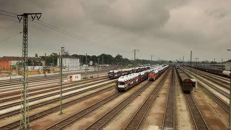 static camera from an elevated view overlooks a train yard in an urban area, multiple trains, tracks, power lines, cargo containers, and buildings under an overcast sky