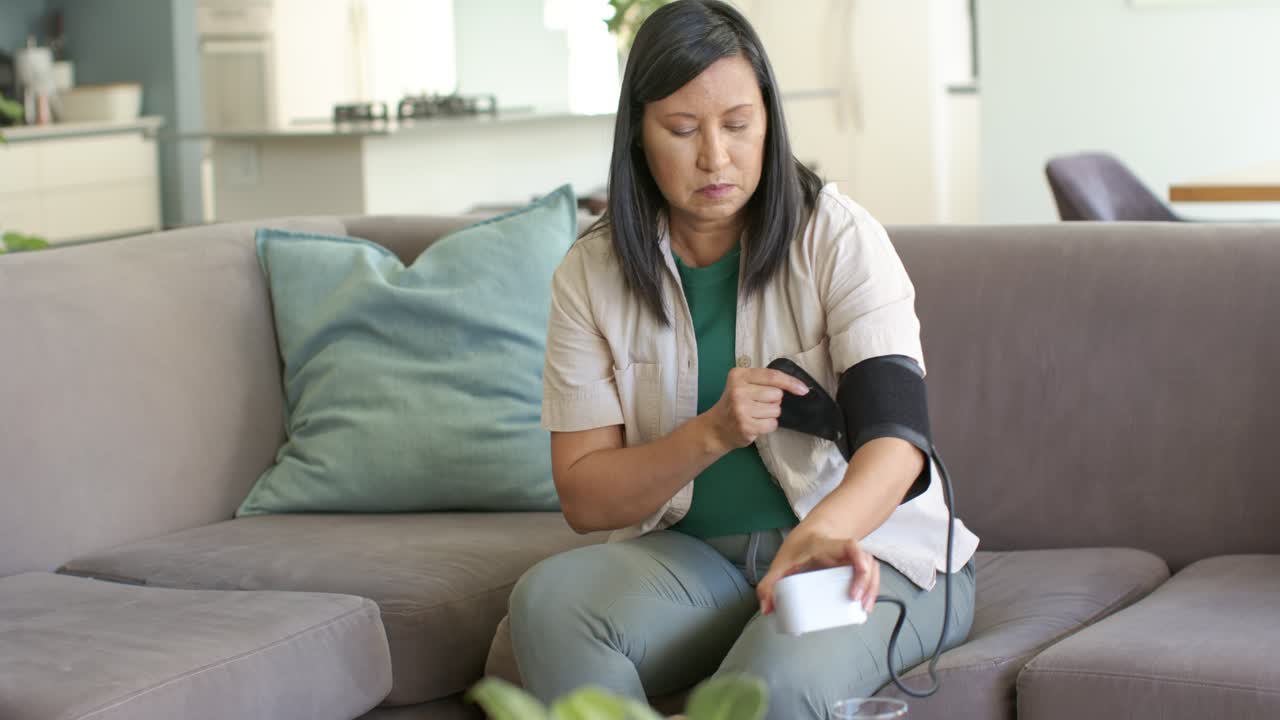 Donna asiatica matura seduta sul divano, che avvolge un bracciale per la pressione sanguigna e regola il monitor per controllare la pressione sanguigna