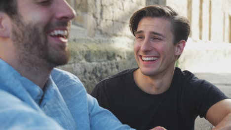 male gay couple sitting outdoors on steps of building laughing together