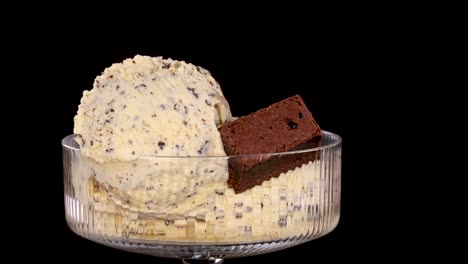 a hand places a chocolate brownie piece onto vanilla ice cream with chocolate chips in a glass bowl, under bright studio lighting against a black background
