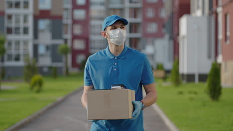 the male deliveryman in a cap and a protective mask and gloves goes with a box in his hands and carries a parcel to the customer. delivering online home orders.