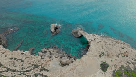 Aerial---The-natural-rock-formation-known-as-the-Bridge-of-Love,-located-in-Ayia-Napa,-Cyprus-clear-turquoise-waters-surround-the-weathered-limestone-arch,-striking-contrast-with-the-rugged-coastline