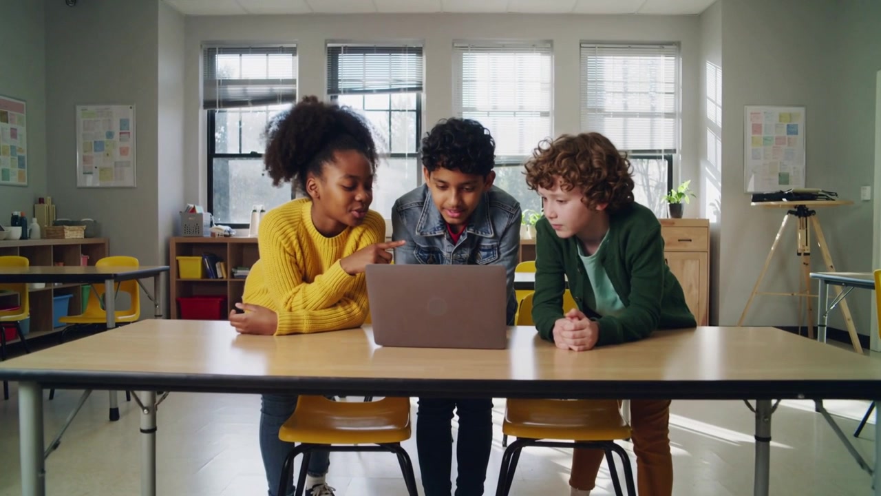Leaning forward, three kids pointing, typing at school desk, using laptop for class task, in yellow