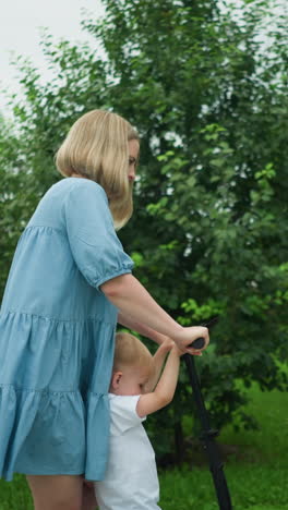 a mother rides a scooter with her young son on a walkway, using her leg to move it forward, her dress flutters gently in the wind