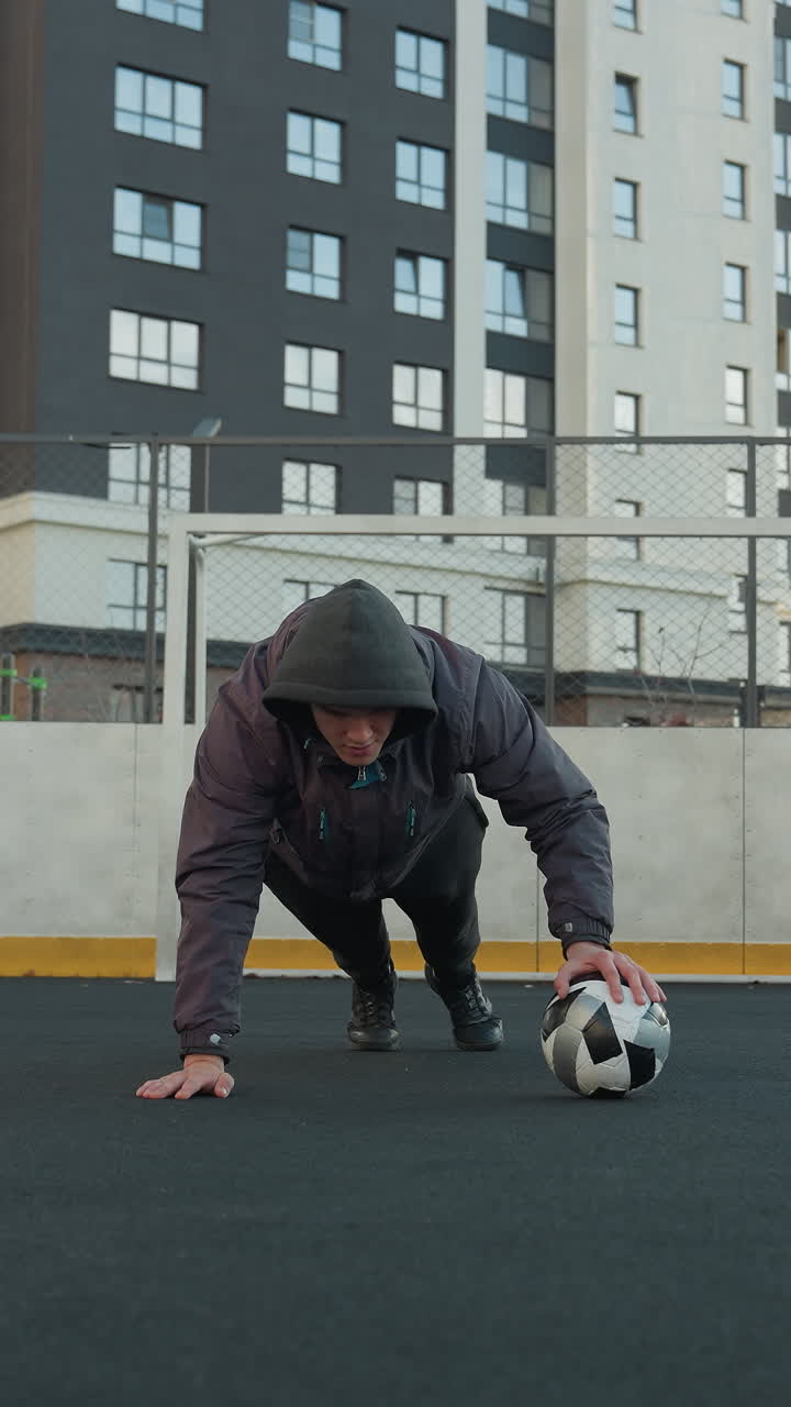Businessman performing push-ups alternating hand placement on soccer ball demonstrating strength, in outdoor urban arena with goal post and residential buildings in the background