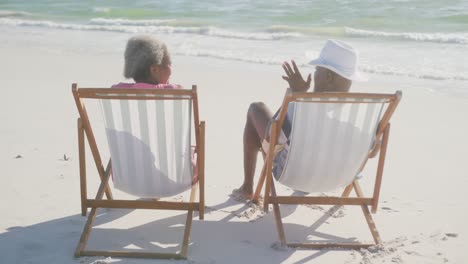 happy senior african american couple sitting on deck chairs at beach, in slow motion