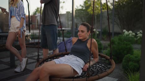 Happy-brunette-girl-in-a-gray-T-shirt-swings-on-a-rope-swing-while-relaxing-with-her-friends-in-a-country-house