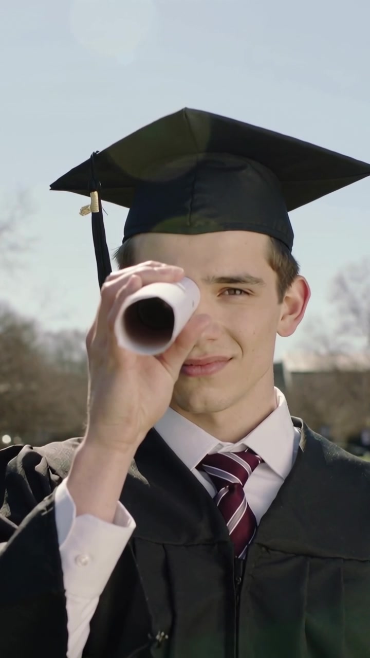 Video verticale: Un laureato con tocco e toga con nappa tiene un diploma arrotolato all'occhio, guardando il campus.
