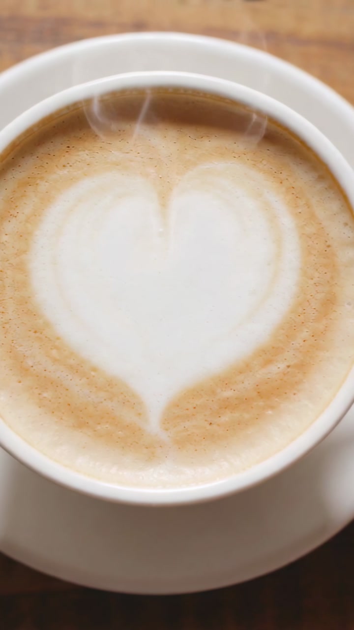 Vertical video: Releasing steam after pouring, white cup showing heart foam on wooden table, saucer