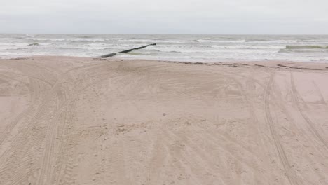 Establishing-aerial-view-of-Baltic-sea-coast-on-a-overcast-day,-old-wooden-pier,-white-sand-beach,-large-storm-waves-crushing-against-the-coast,-climate-changes,-wide-drone-shot-moving-forward-low