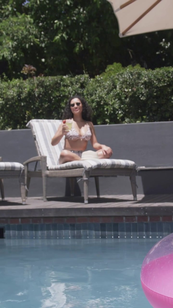 Vertical video: Sun warming, woman in patterned bikini sipping cup on pool lounge with pink float