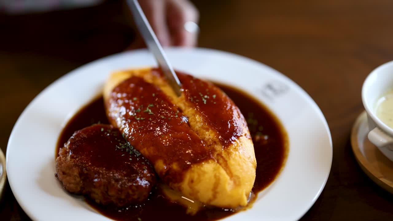 Hand slices omurice with demi-glace beside hamburg steak, warm lighting, close-up, steady camera
