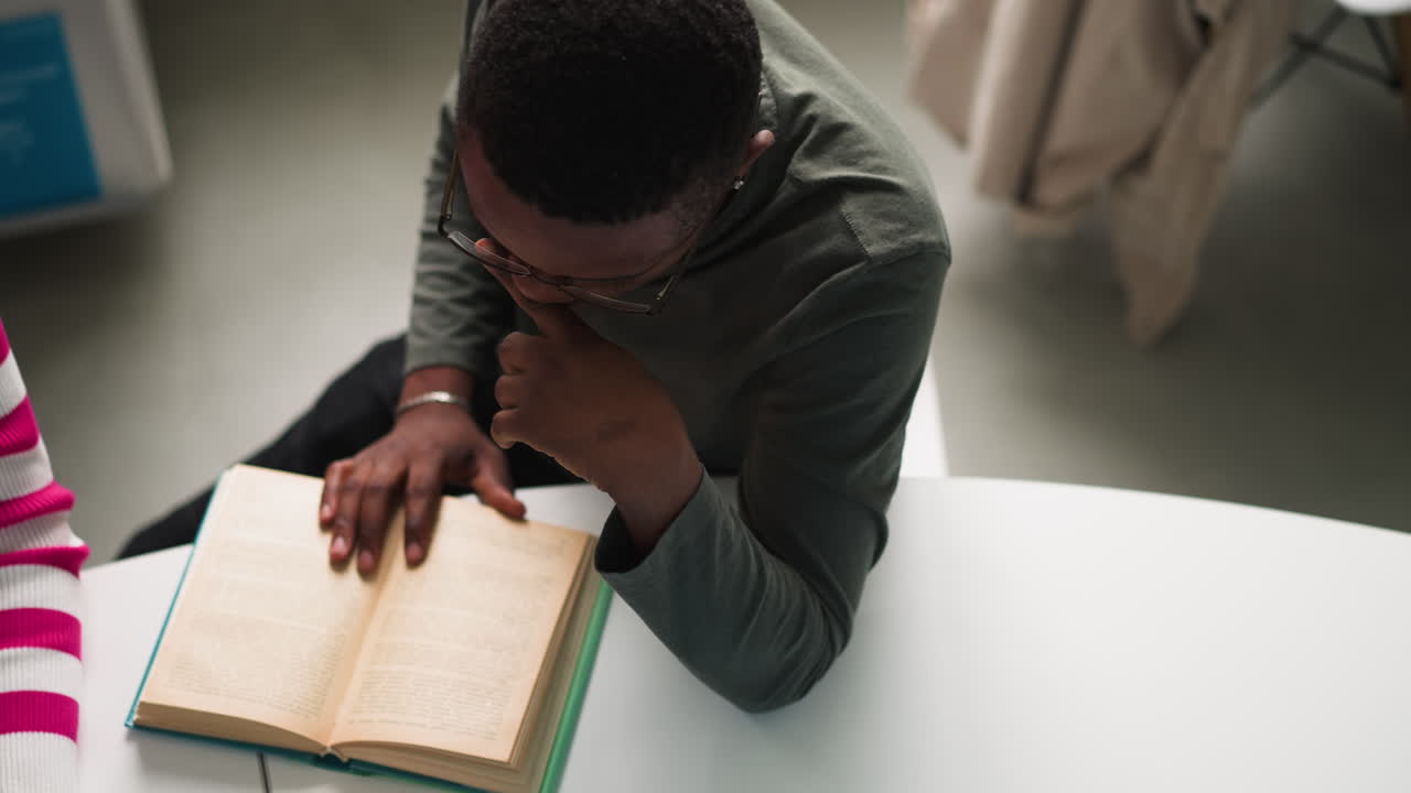 Black man reads book sitting at table upper view. Student studies textbook doing graduate work in reading hall. African American guy in library