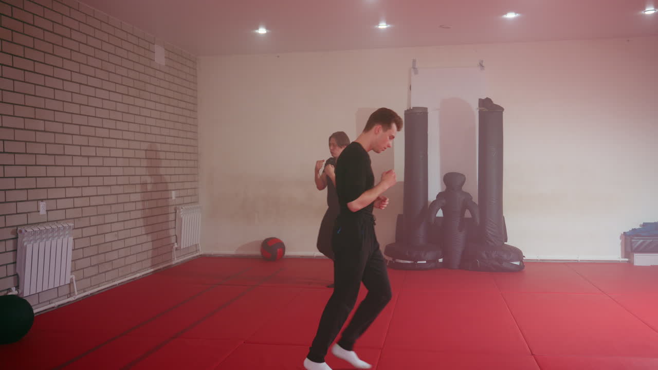 Two athletes in black sportswear train inside martial arts gym on red mat, one practicing boxing stance while other jogs, focusing on endurance,discipline, and preparation for combat training