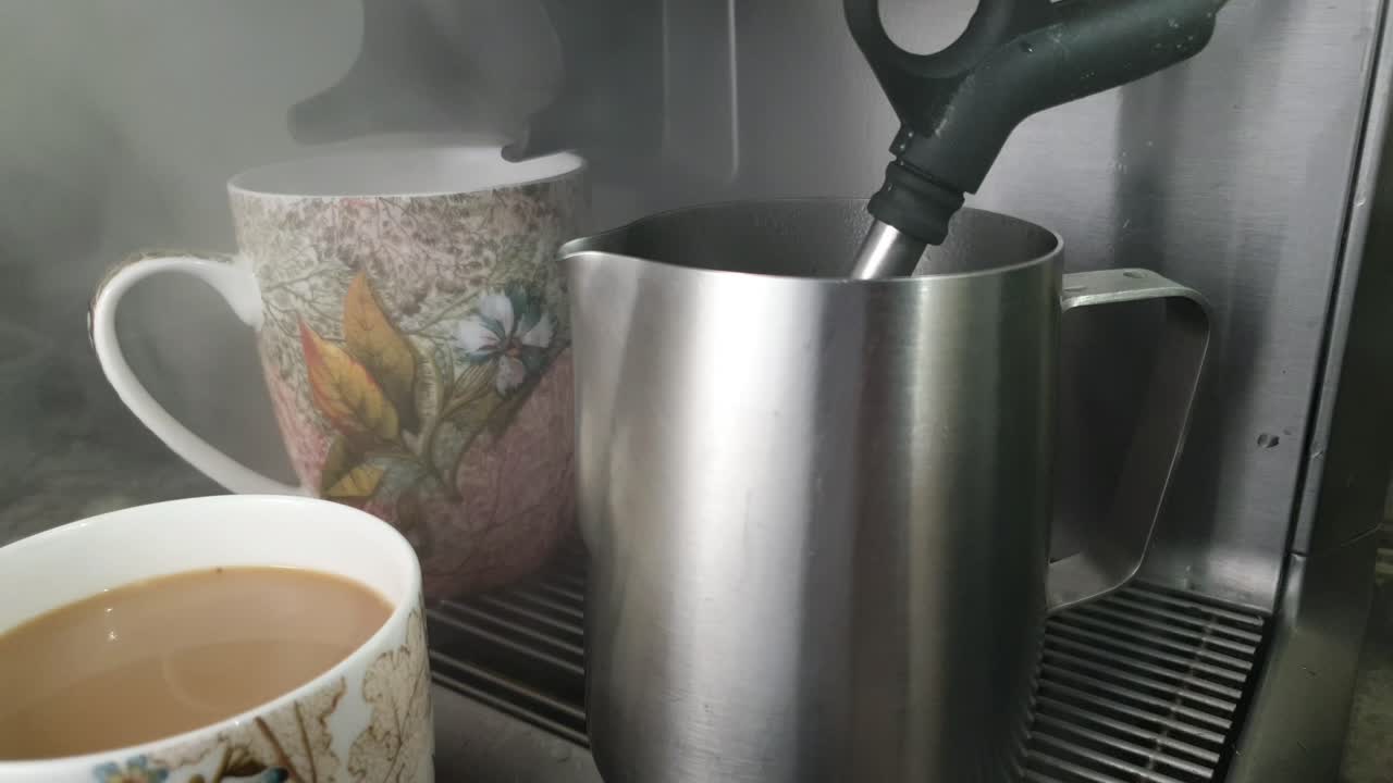 el capuchino se prepara con una máquina de café mientras se humea espuma de leche en una taza de acero inoxidable, dos hermosas tazas de café con motivos florales esperan a un lado la dosis de cafeína