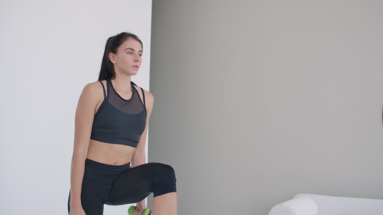 A young brunette woman performs concentrated pacing on a chair in a bright interior apartment with dumbbells in her hands.