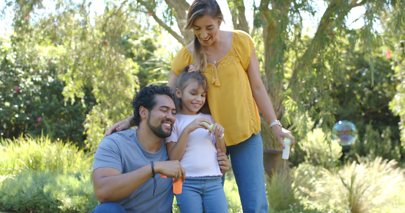 Família diversa brincando com varinhas de bolhas de sabão no jardim, com o pai apontando, a filha soprando e a mãe se juntando.
