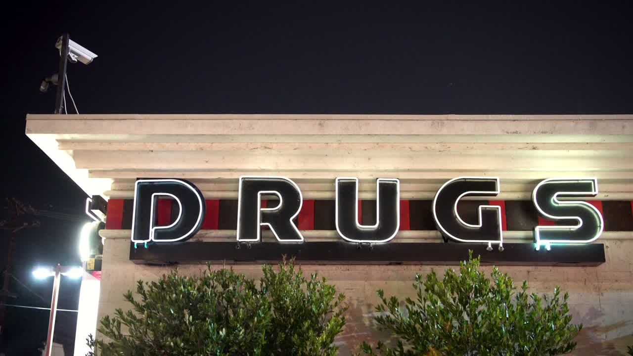 Illuminated neon sign saying DRUGS at night on a building's side.