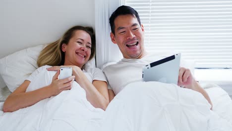 침대에 휴대 전화 디지털 태블릿을 사용하는 행복한 커플