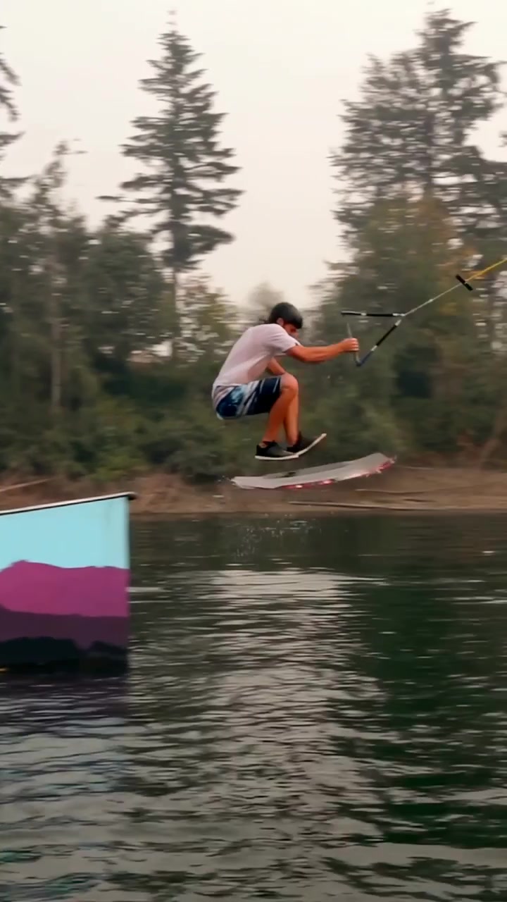 Video verticale: Wakeboarder che taglia l'acqua, tiene la corda, tirato da un cavo sul lago, cavalca il kicker e cade.