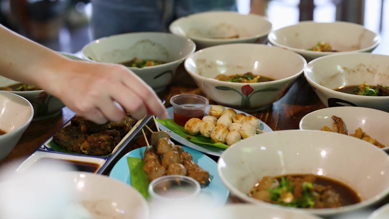 Tampilan dekat tangan menjangkau bakso bakar dan mangkuk sup mi di atas meja kayu.