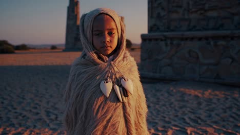youthful child wearing traditional garments and textured fur cloak with shell pendants, standing silently amid desert landscape, ancient stone ruins illuminated by golden sunset light