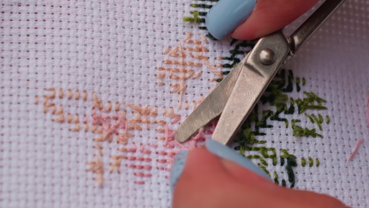 Extreme close up craft person cutting thread on embroidered fabric using metal scissors, blue nails visible while creating precise detail, showing patience and artistry