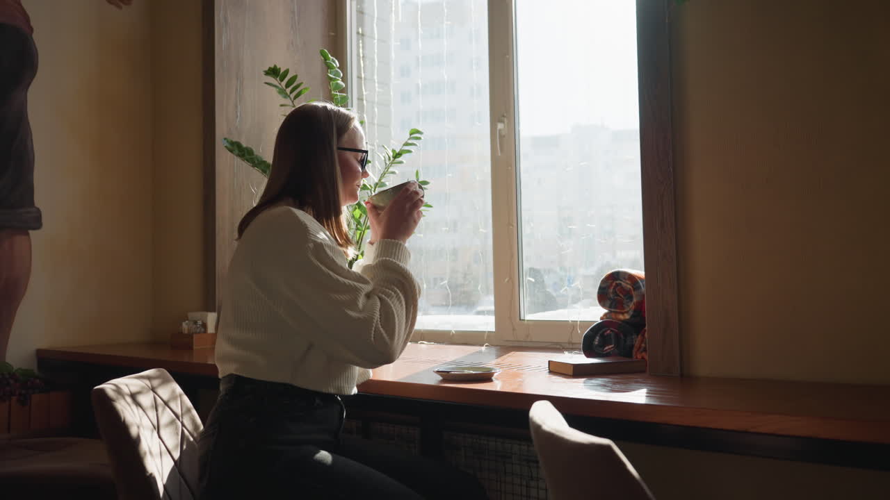 Young girl drinks tea beside window as sunlight streams in, creating dreamy effect, potted plant adds calm touch while distant cityscape shows through glass in quiet, thoughtful atmosphere