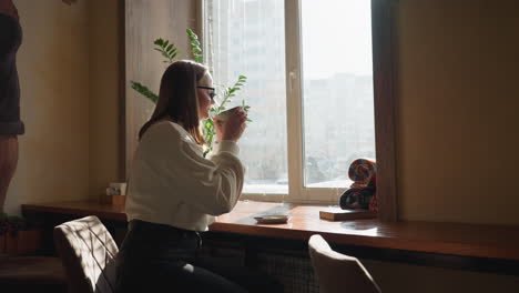 young girl drinks tea beside window as sunlight streams in, creating dreamy effect, potted plant adds calm touch while distant cityscape shows through glass in quiet, thoughtful atmosphere