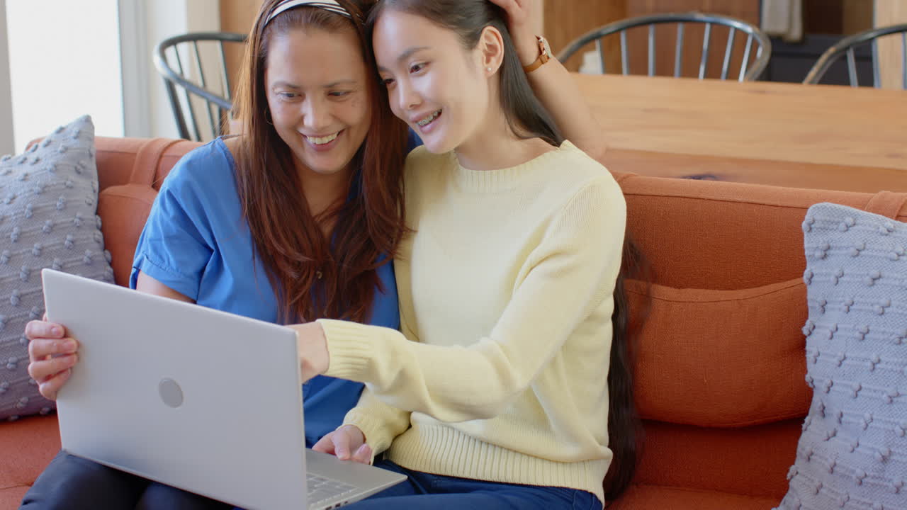 Smiling asian mother and daughter sitting on couch using laptop together at home