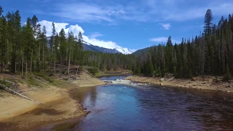 Niedrige-Luftaufnahme,-Die-An-Einem-Hellen,-Sonnigen-Nachmittagsfrühlingstag-In-Der-Waldwildnis-Colorados-Mit-Majestätischen-Schneebedeckten-Bergen,-Blauem-Himmel-Und-Wolken-Langsam-Den-Breiten,-Gewundenen-Fluss-über-Eine-Große-Kaskade-Hinauffliegt