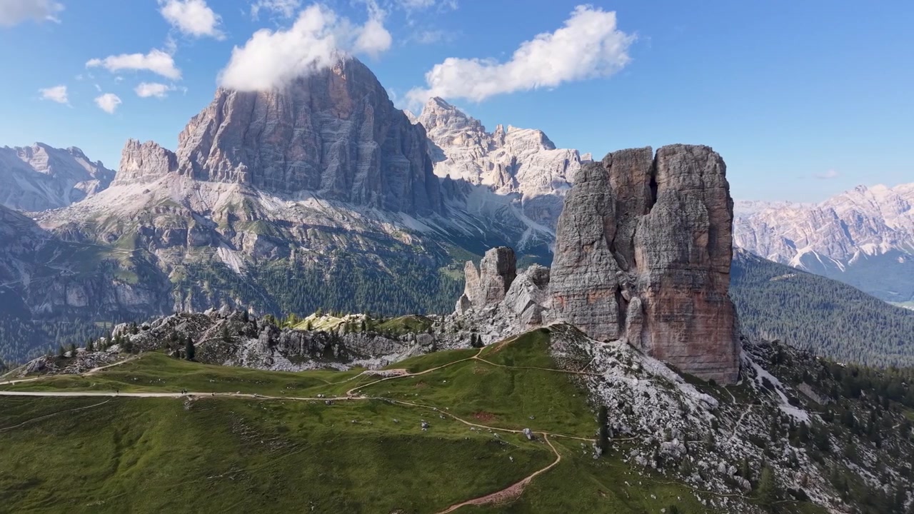 Vista aerea con drone delle formazioni rocciose delle Cinque Torri con l'imponente massiccio delle Tofane sullo sfondo, circondate da prati alpini, pendii rocciosi, sentieri escursionistici e nuvole sparse.