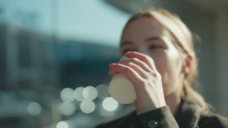 Close up of lady with warm smile drinking soda while walking past glass building reflecting urban surroundings, with sunlight highlighting her face and creating soft background bokeh